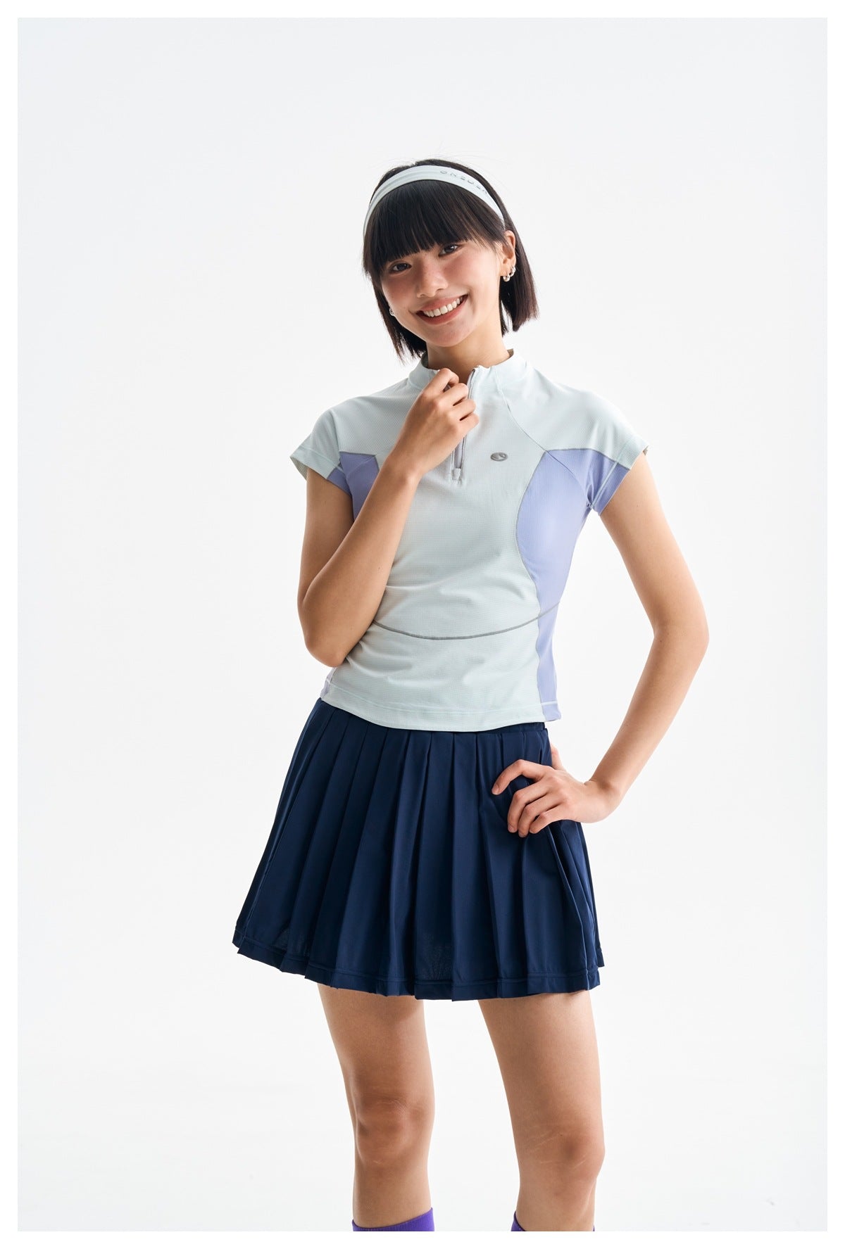 Woman in a pale blue quarter-zip top with lavender side panels and a navy pleated skirt, smiling with one hand at the collar.