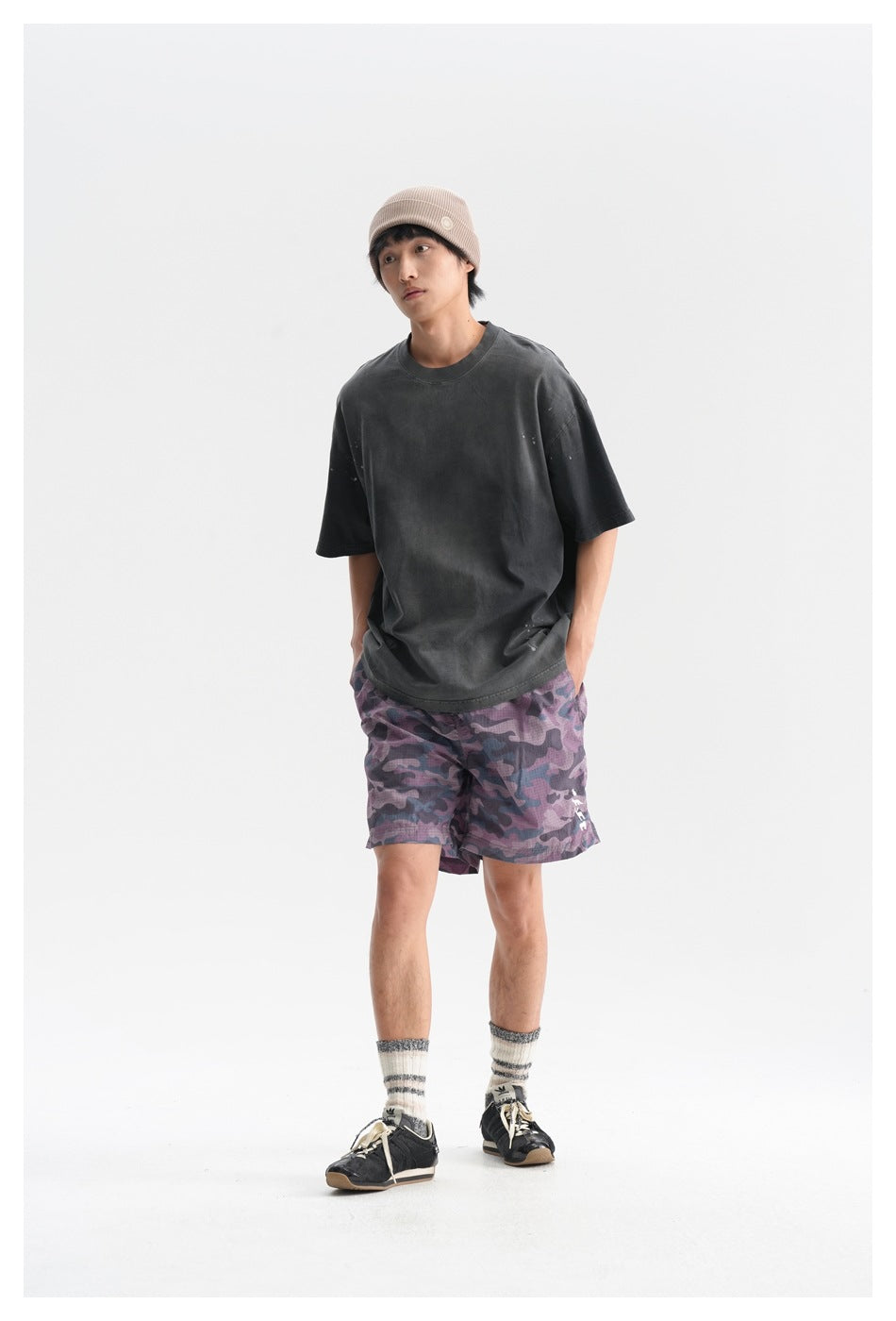Model wearing a charcoal oversized T-shirt and purple camouflage shorts, posed at a slight angle with hands in pockets.