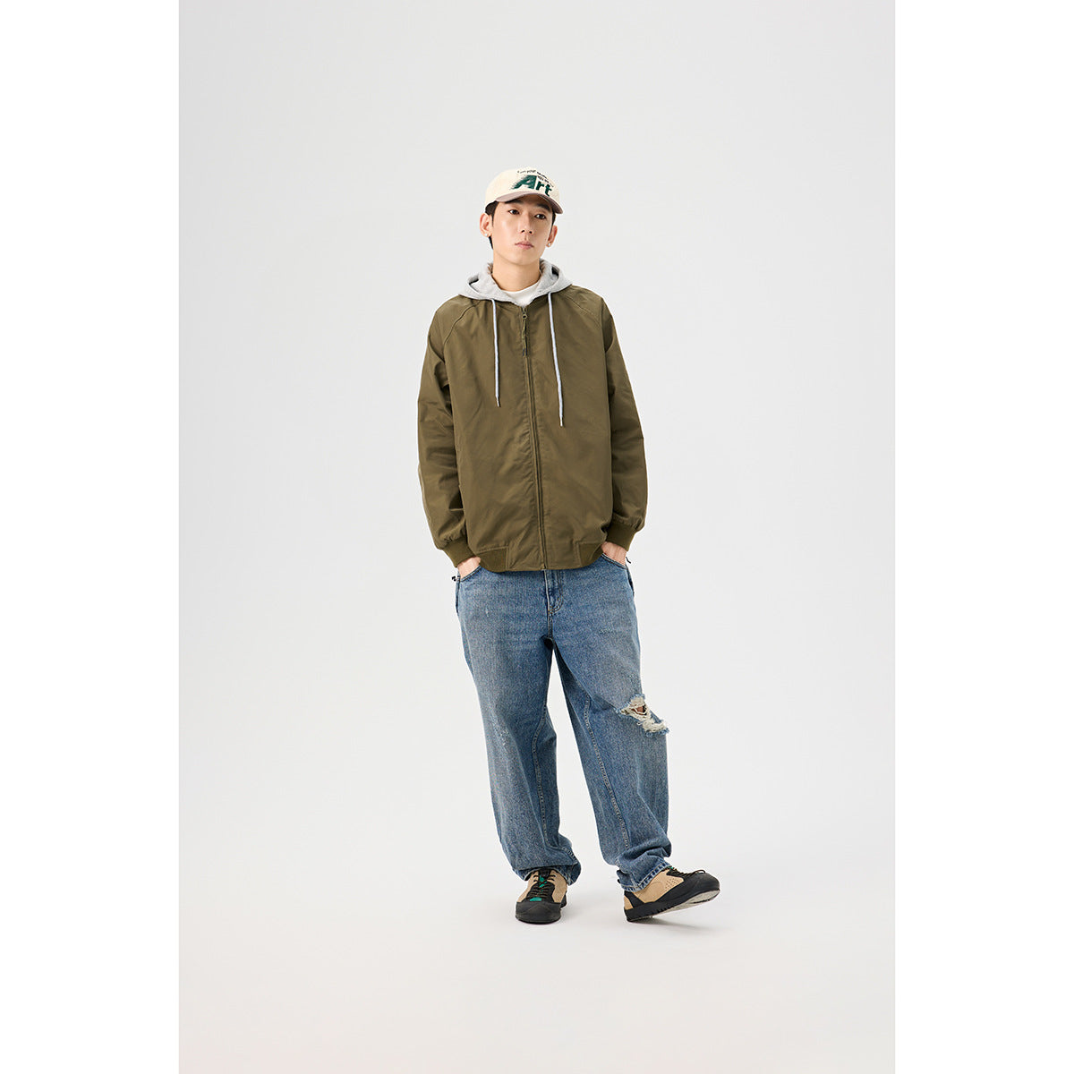 Model wearing a zipped olive jacket with a gray hood, loose blue jeans, and a beige cap.