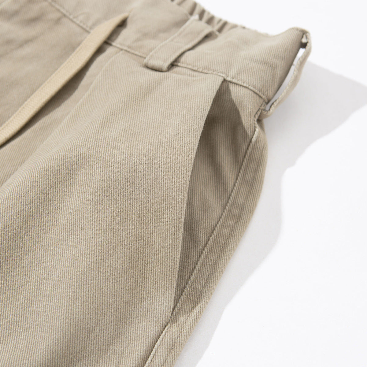 Angled close-up of beige trousers showing a belt loop, waistband, side seam and drawstring.