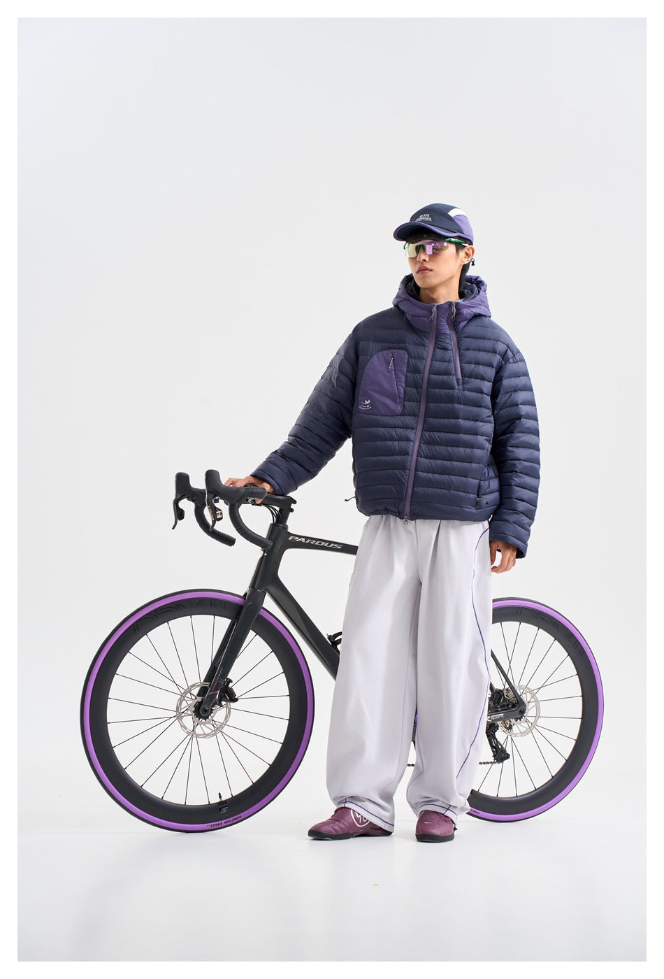Full-length front view of a person wearing a navy quilted jacket with a purple hood and white trousers beside a bicycle.