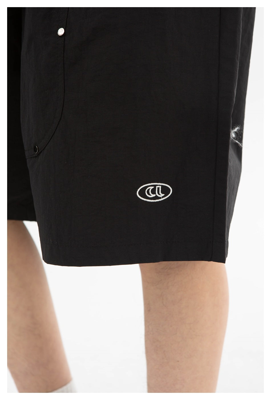 Close-up of black shorts showing a small white oval emblem, a snap-fastened panel, and the hem.