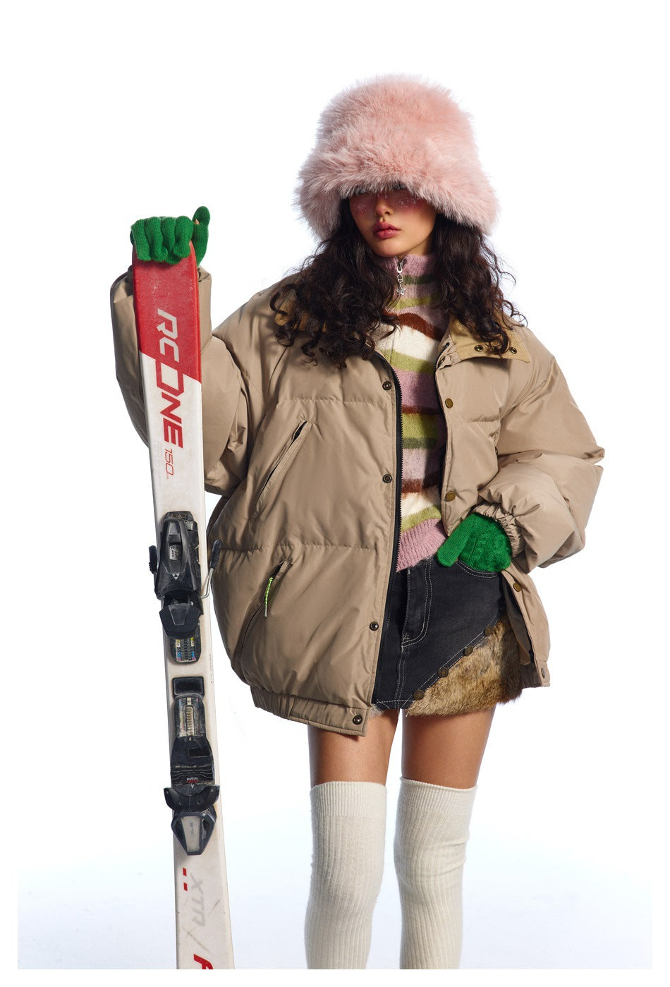 Model wearing an oversized beige puffer jacket with a striped sweater, pink fur hat and green gloves, holding skis.