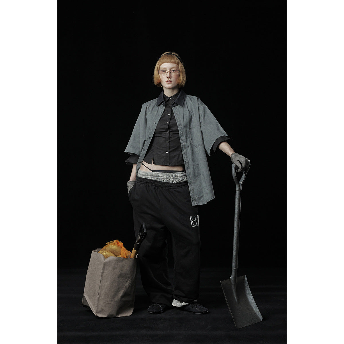 Model wearing a loose gray short-sleeve button-up shirt over black layers and black trousers against a dark background.