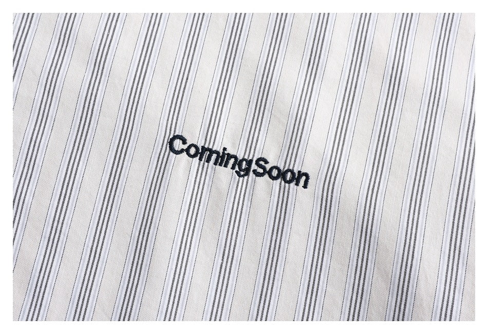 Close-up of gray pinstripes and black “Coming Soon” embroidery on white shirt fabric.