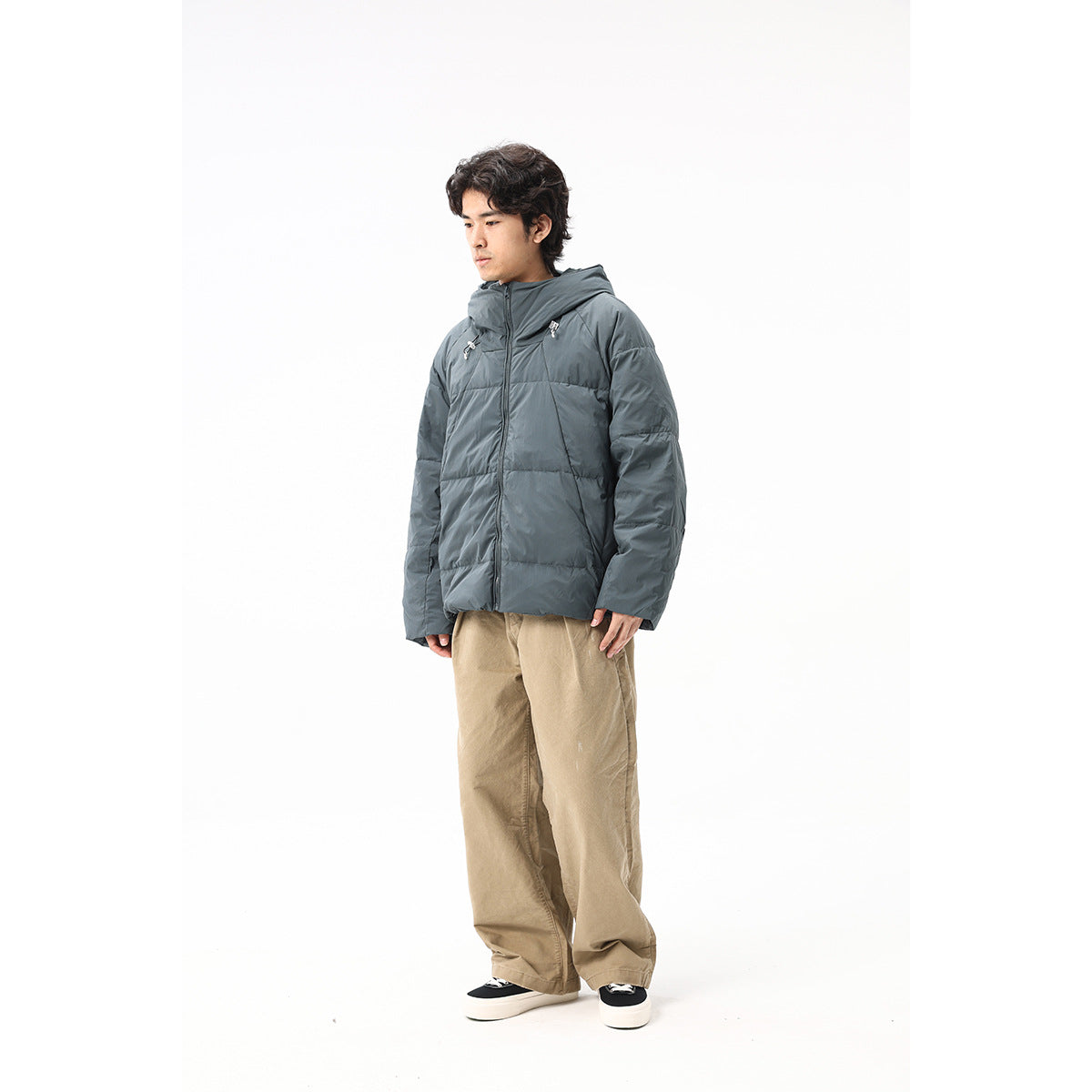 Full-length model wearing a slate-gray hooded puffer jacket with tan trousers, shown from a three-quarter front angle.