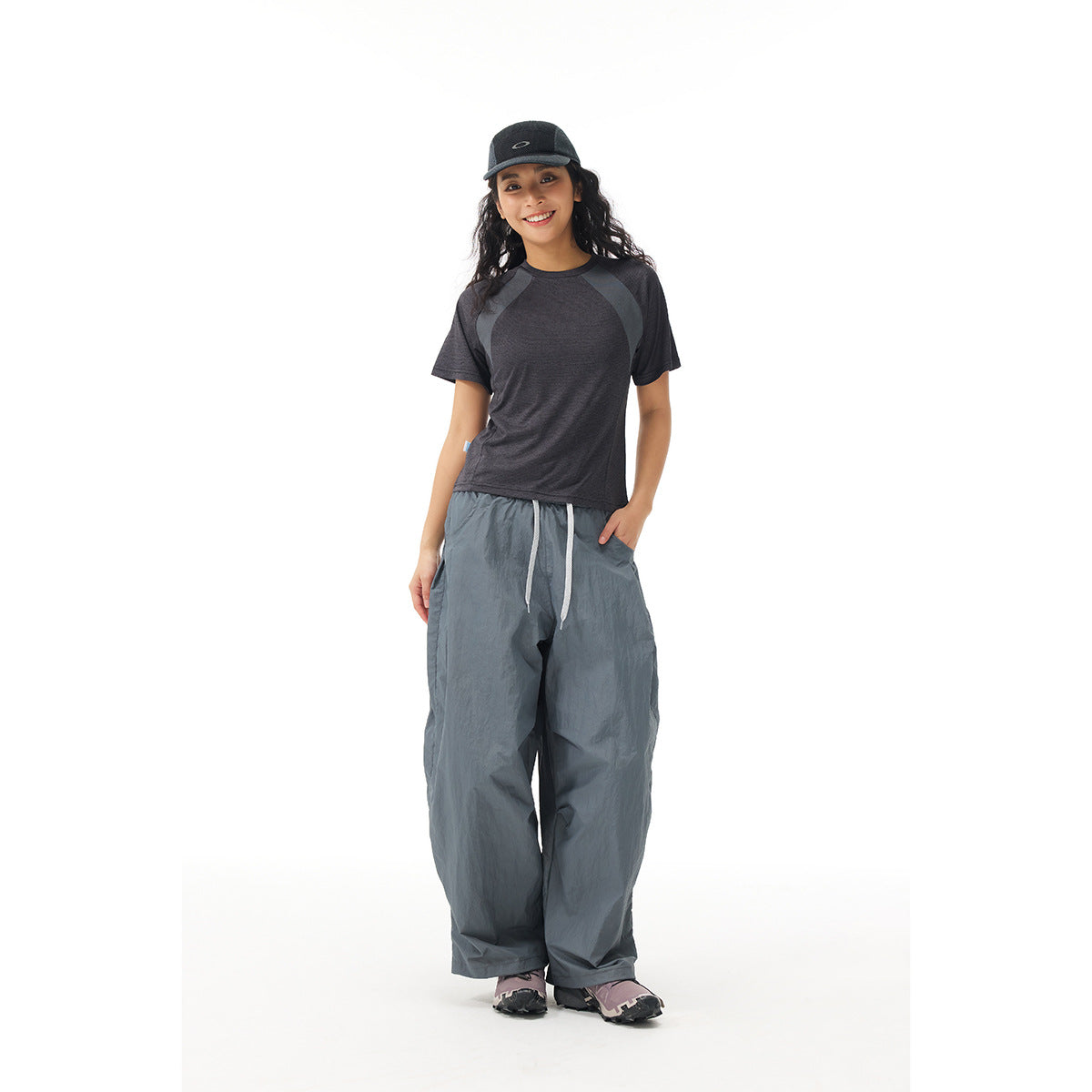 Model wearing a charcoal color-block T-shirt, loose slate-blue drawstring pants, and a black cap.