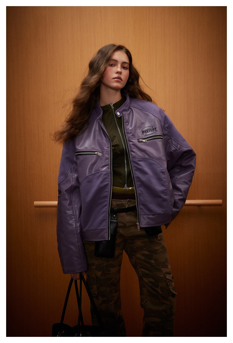 Model wearing a purple zip-front jacket with chest zippers and camouflage trousers against a wood-paneled wall.