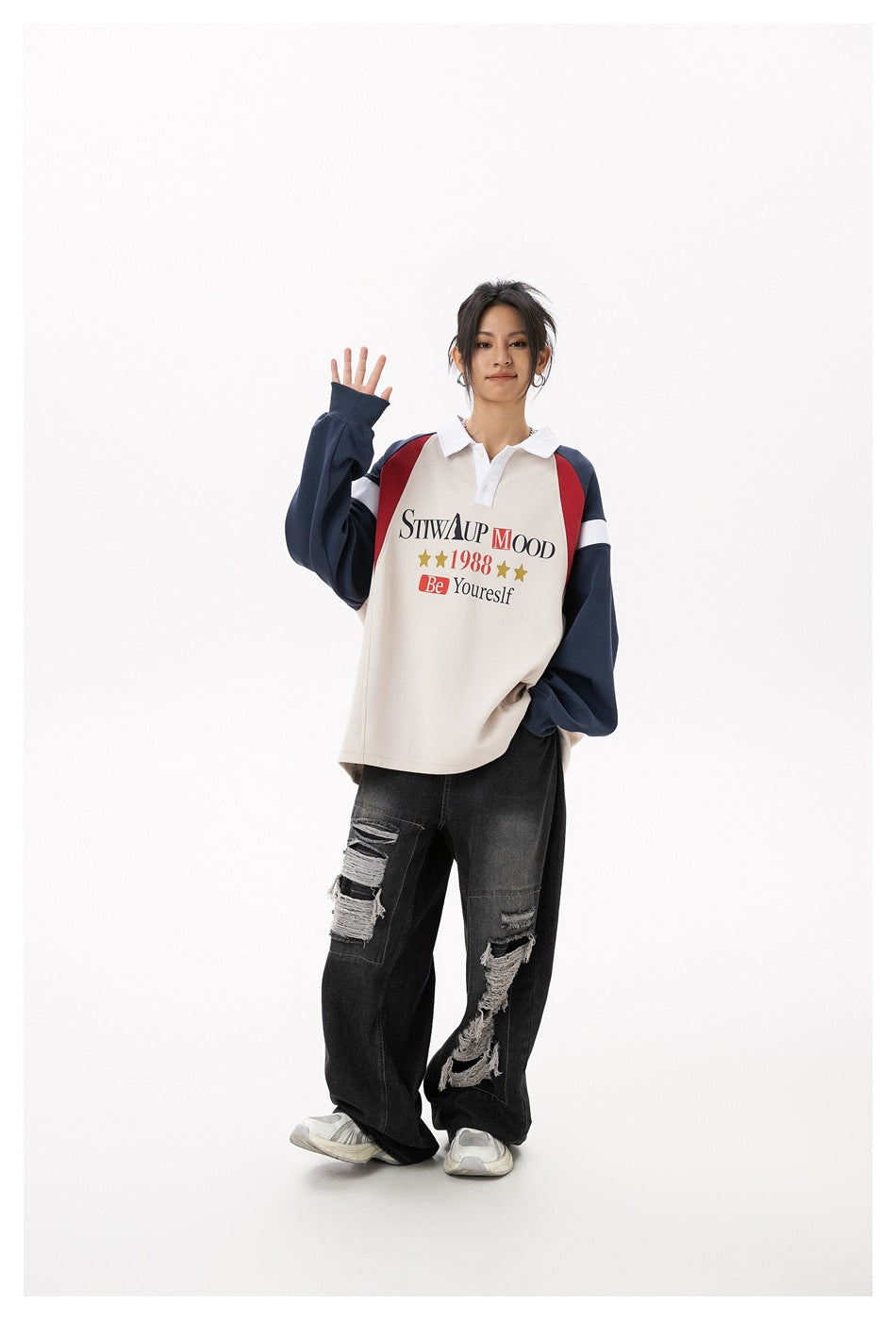 Full-length front view of a model waving in a cream polo sweatshirt with navy and red panels and loose black ripped jeans.