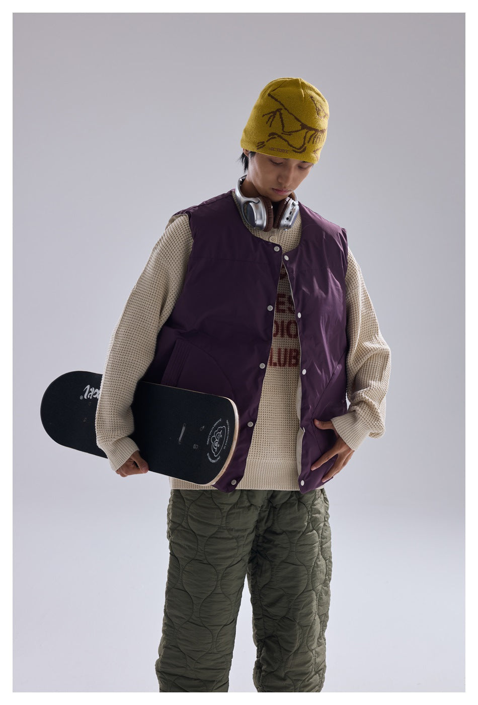 Model wears a plum snap-front vest over a cream sweater with olive quilted trousers and holds a skateboard.