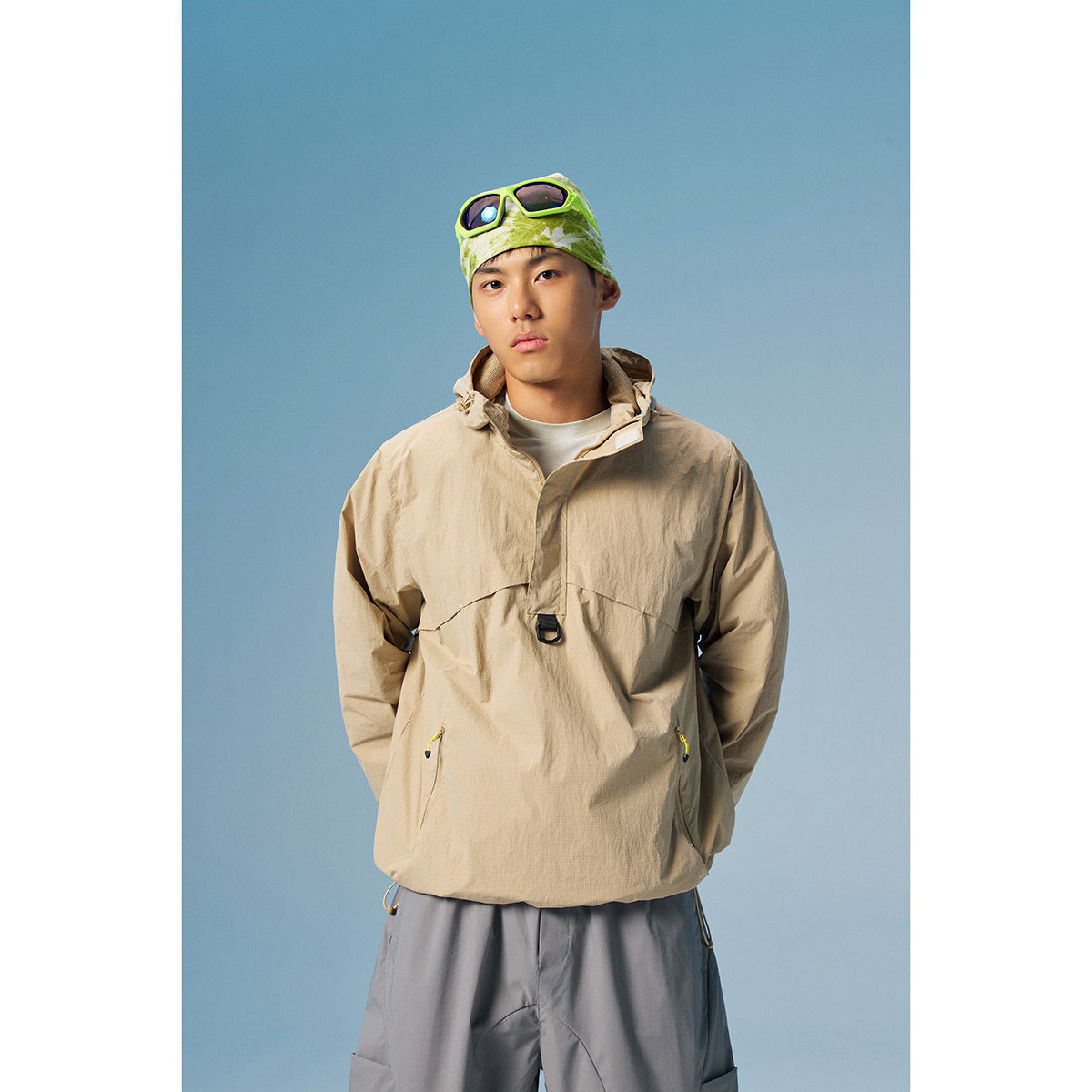 Model wearing a beige hooded pullover jacket with yellow zipper pulls and gray trousers.
