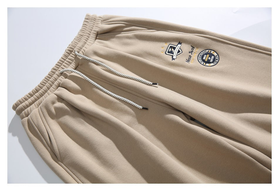 Angled close view of beige jogger pants with an elastic waist, braided drawstrings, and embroidered chest-style patches.