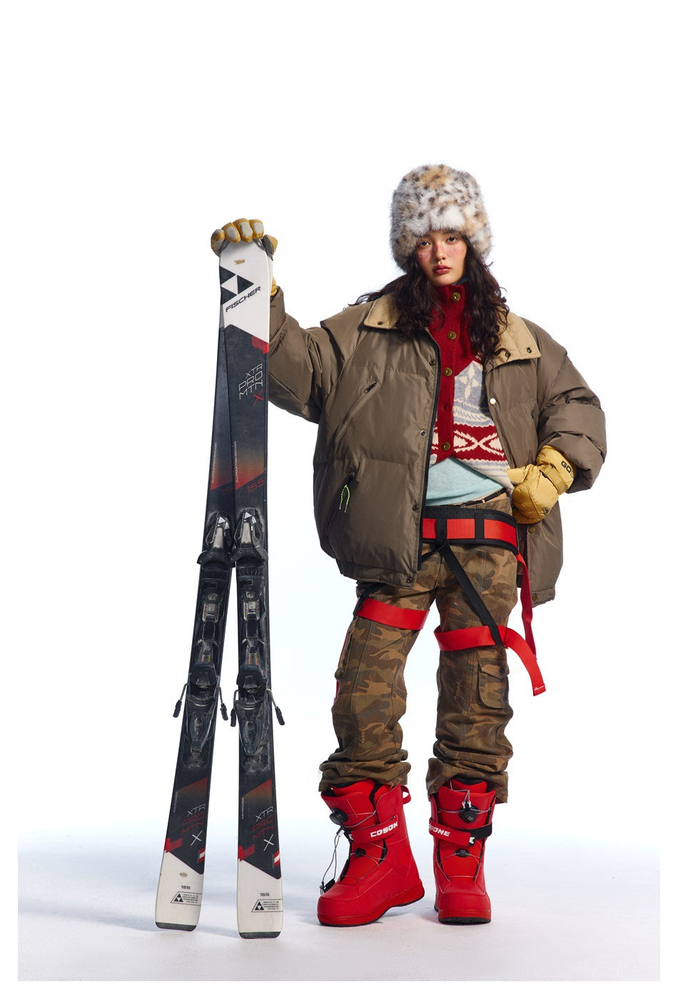 Model wearing a dark taupe oversized puffer jacket over a patterned sweater, with camouflage trousers and a spotted fur hat.