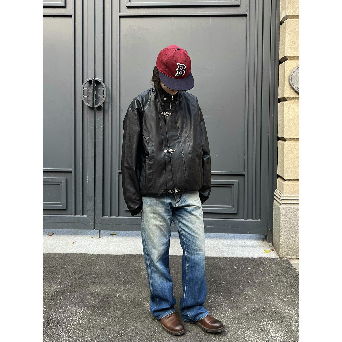 Person wearing black stand-collar jacket with metal hook clasps, loose blue jeans, brown shoes and red cap by a gray door