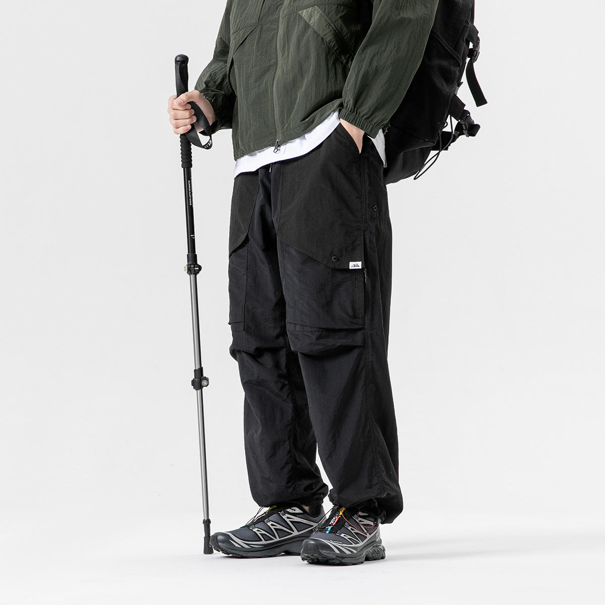 Side view of a model wearing black cargo pants with large flap pockets, paired with a green jacket and hiking gear.
