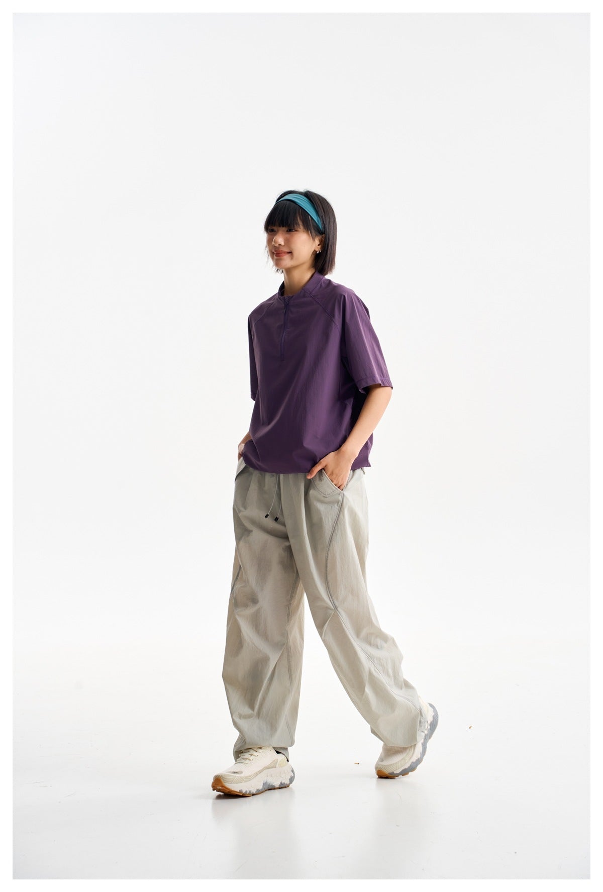 Model wearing a purple short-sleeve quarter-zip top and loose light gray trousers, shown against a white studio background.