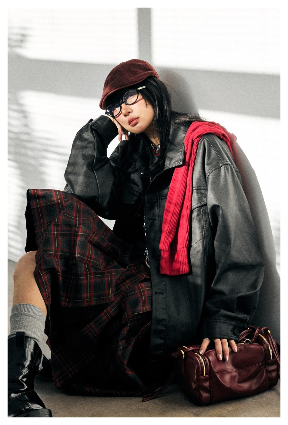 Model seated in an oversized black jacket, with a red scarf and red-and-black plaid skirt.