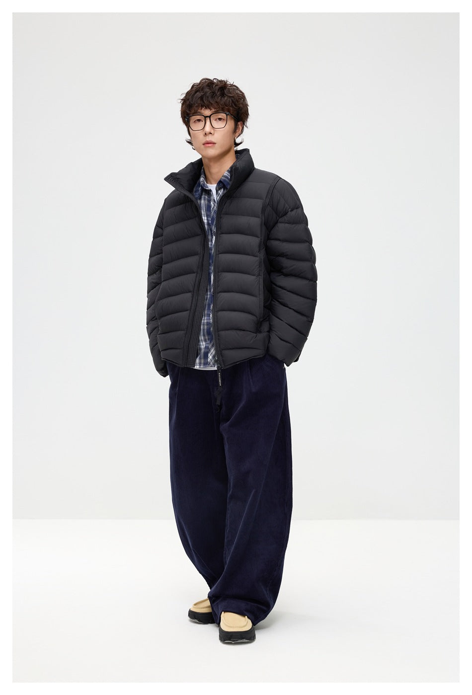 Full-length model wearing a black quilted puffer jacket open over a blue plaid shirt and navy wide-leg trousers.