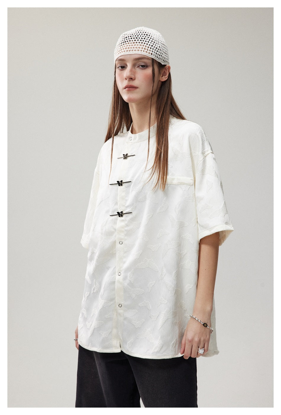 Model wearing an oversized white patterned shirt with short sleeves, a chest pocket and decorative frog closures.