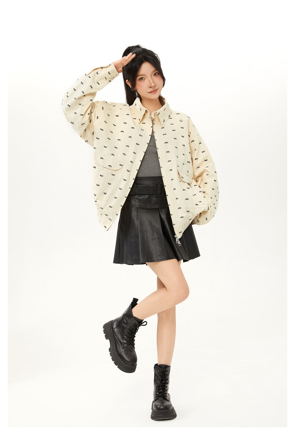 Model in a bow-print apricot jacket posing with one hand raised, wearing a gray top, black skirt and black boots