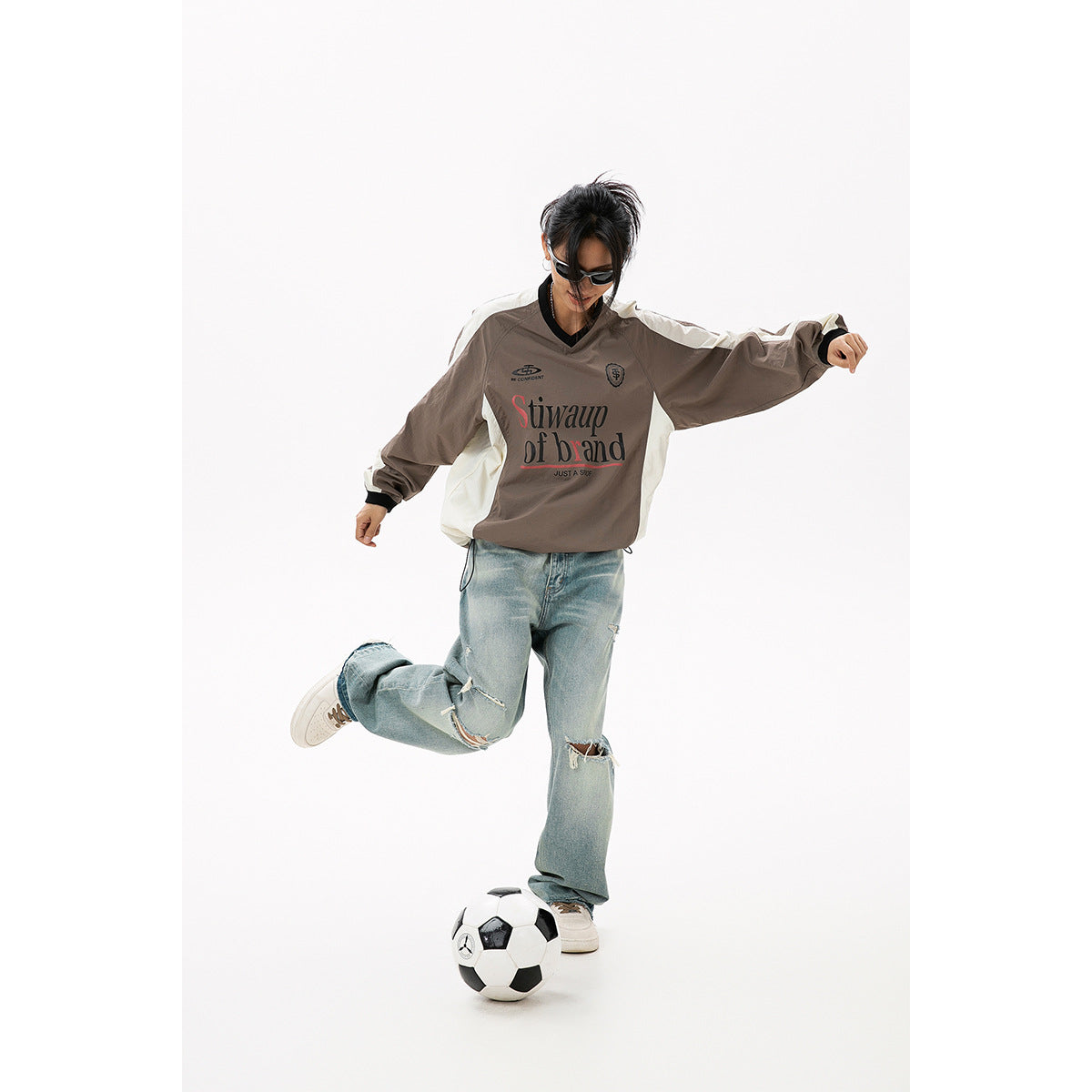 Model wearing a taupe V-neck pullover with cream side panels and black trim, posed with a soccer ball.