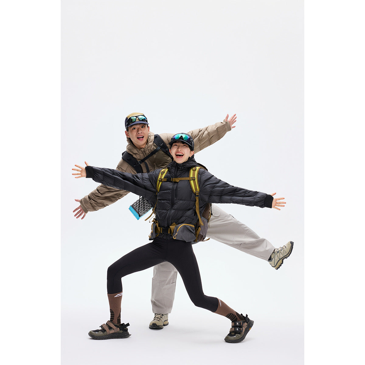 Two hikers pose in black and taupe hooded puffer jackets with backpacks against a plain white studio background.
