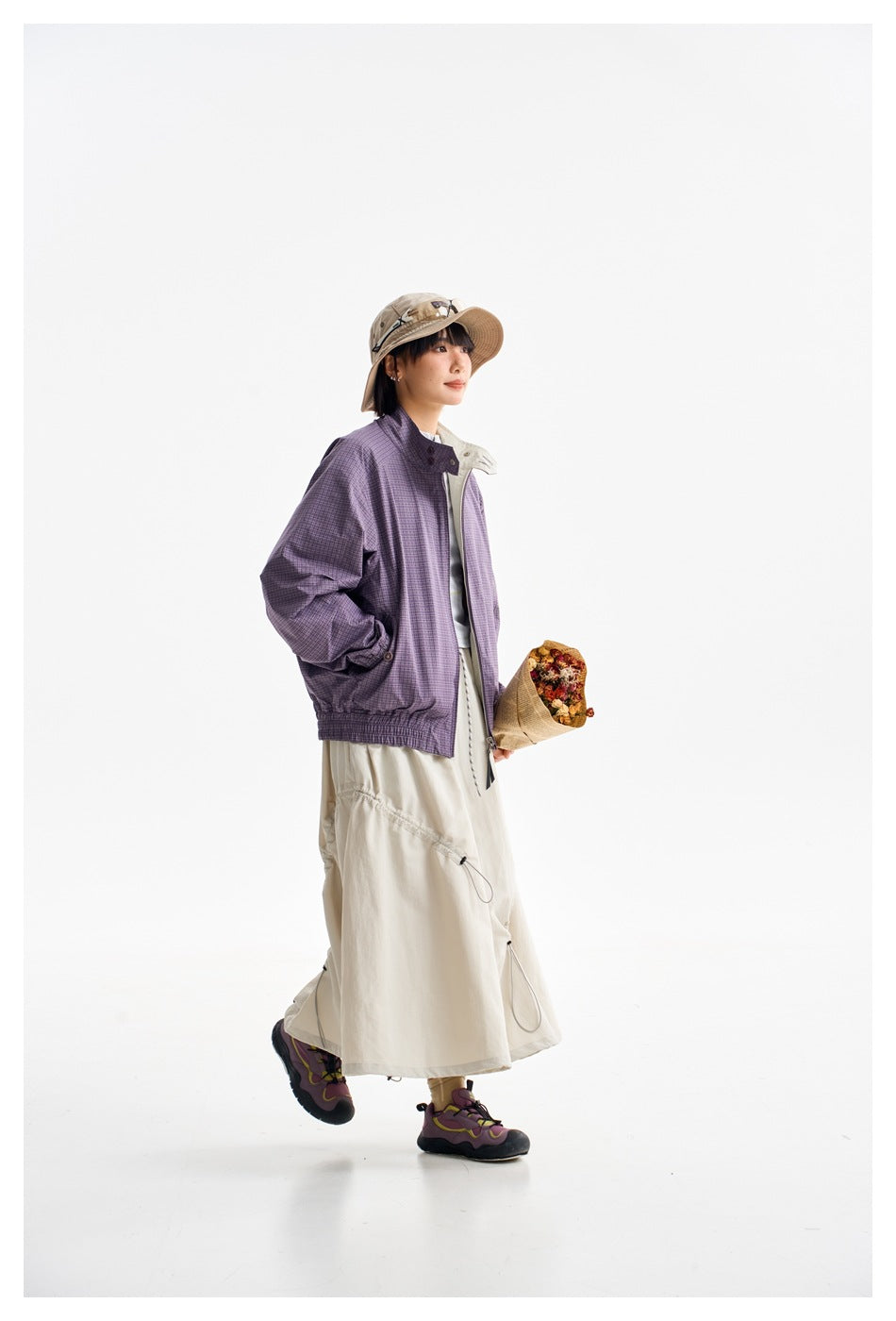 Side view of a model wearing a purple plaid jacket and cream skirt, holding a small bouquet.