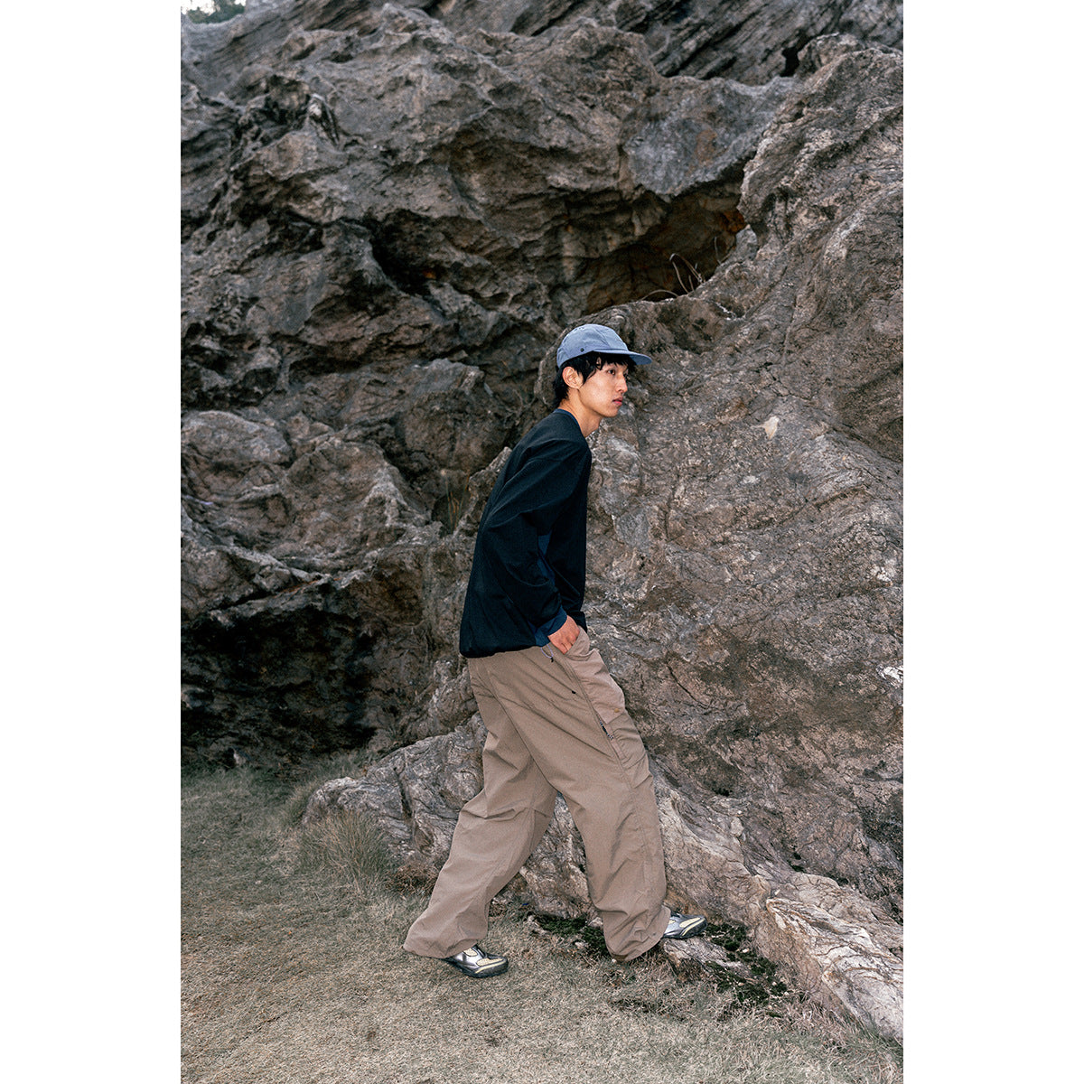 Model in a black long-sleeve shirt, loose taupe trousers, and blue cap beside a rocky cliff.