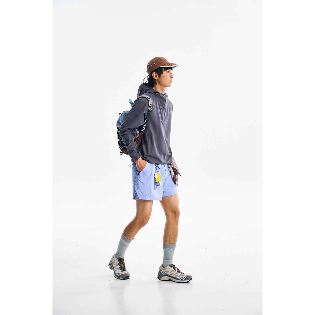 Model wearing light blue shorts with a dark hooded top, cap and backpack against a white background.