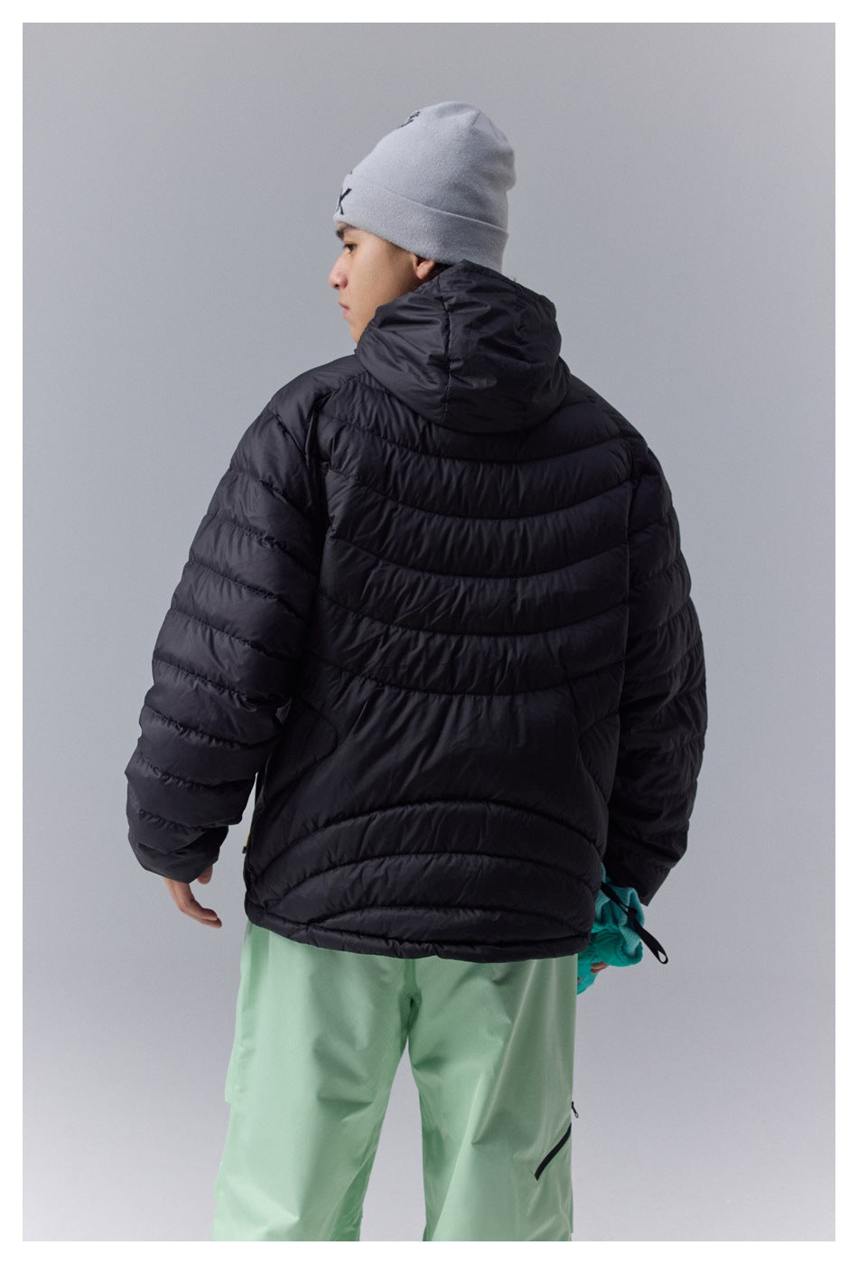 Back view of a model wearing a black hooded quilted jacket and mint-green snow pants.