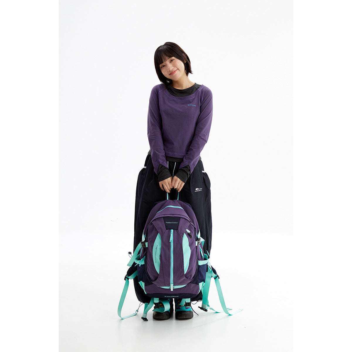 Model wearing a purple long-sleeve top over a black shirt, holding a purple and turquoise hiking backpack.