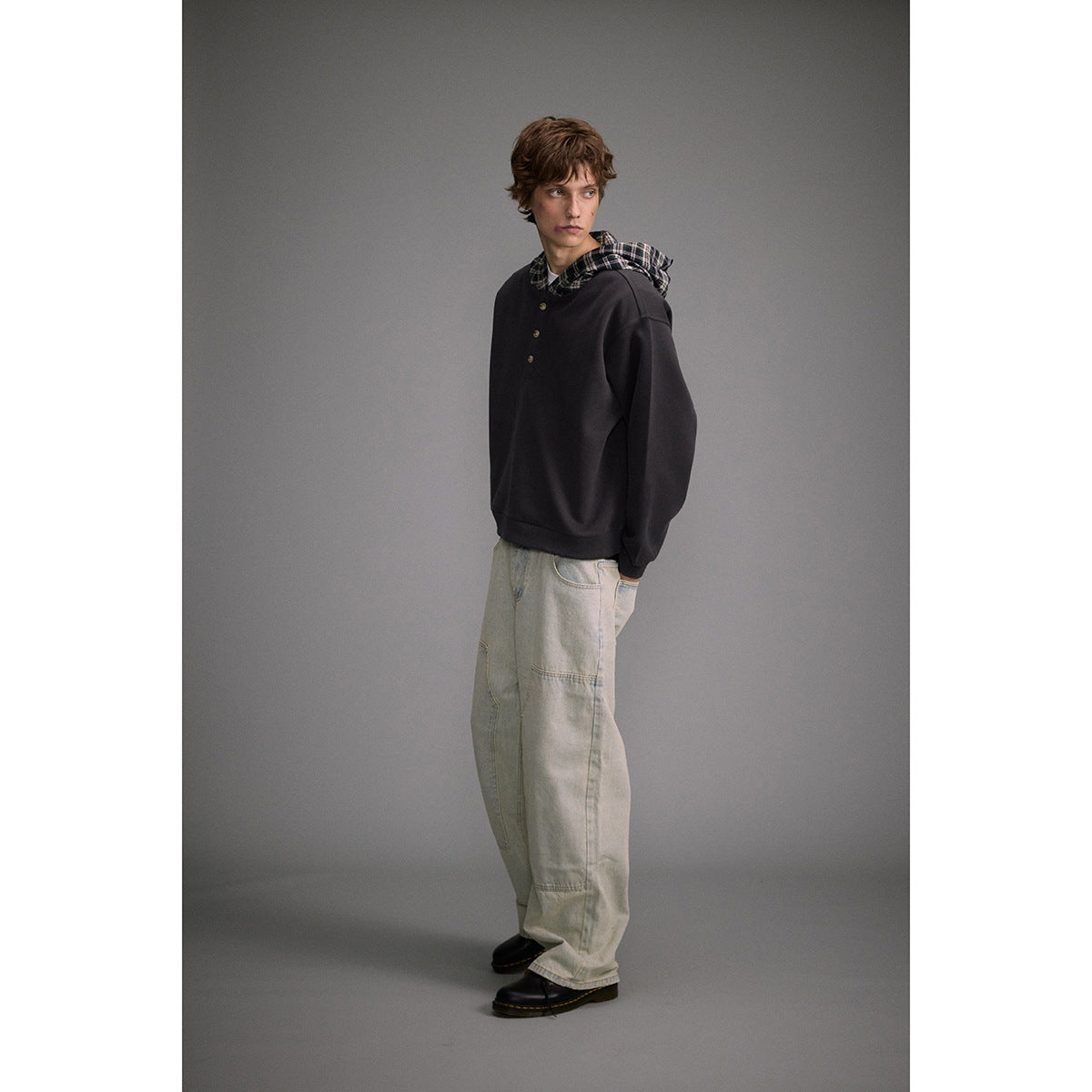 Full-length view of a man wearing a charcoal plaid-lined hooded pullover and loose light-wash jeans.