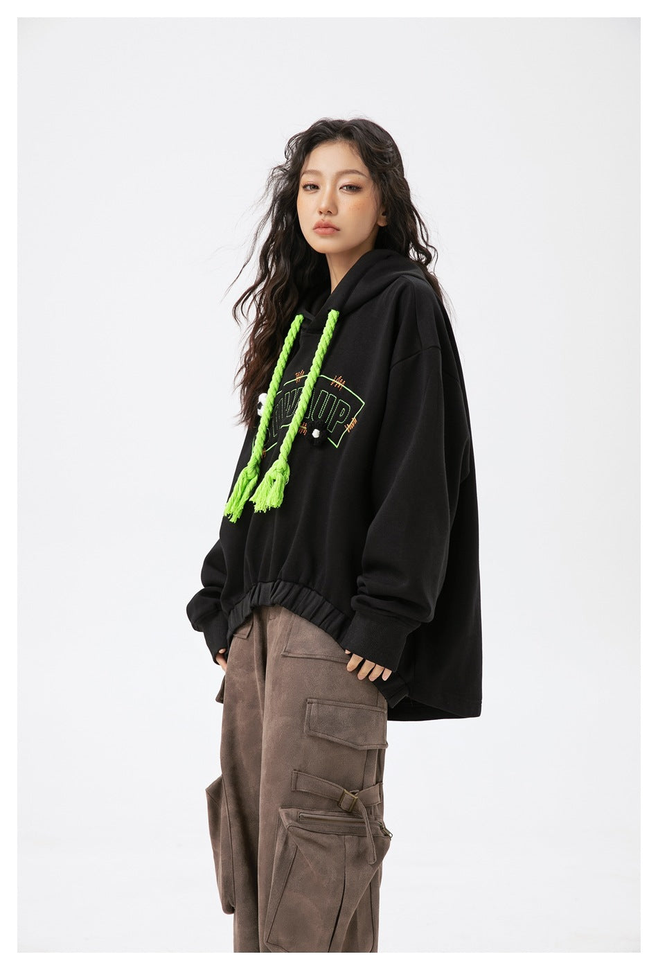 Model in a black oversized hoodie with neon green rope drawstrings and brown cargo pants, shown from a three-quarter side
