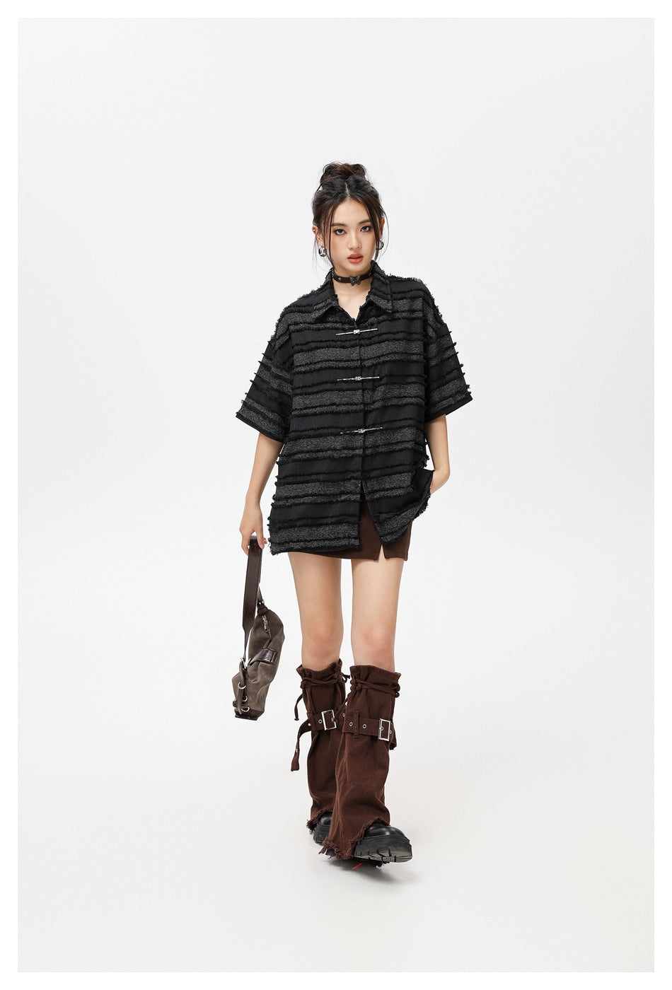 Model walking in an oversized black striped shirt with metal clasps, brown mini skirt, and tall brown boots.
