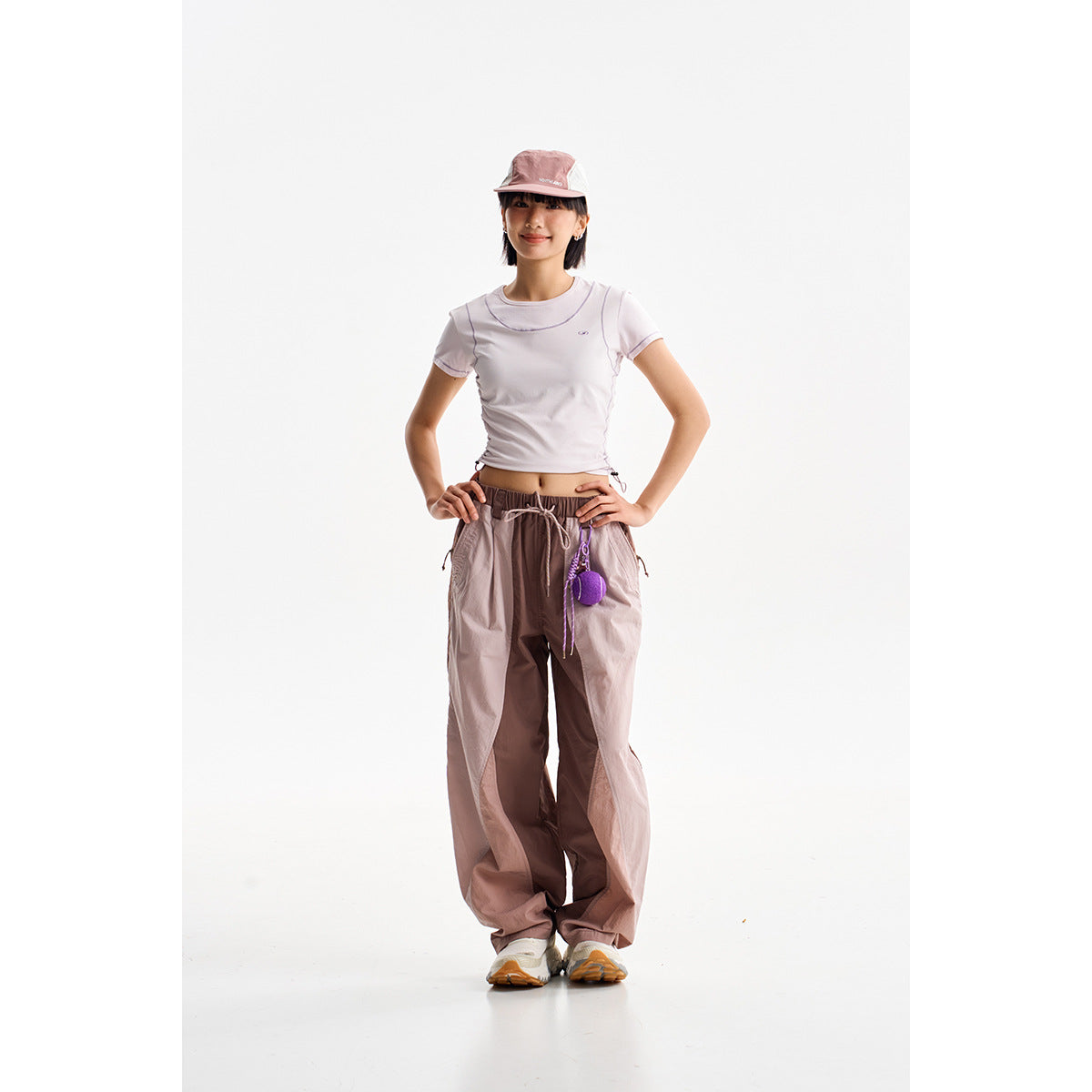 Model wearing a pale pink short-sleeve top and cap with loose taupe drawstring pants and white sneakers.