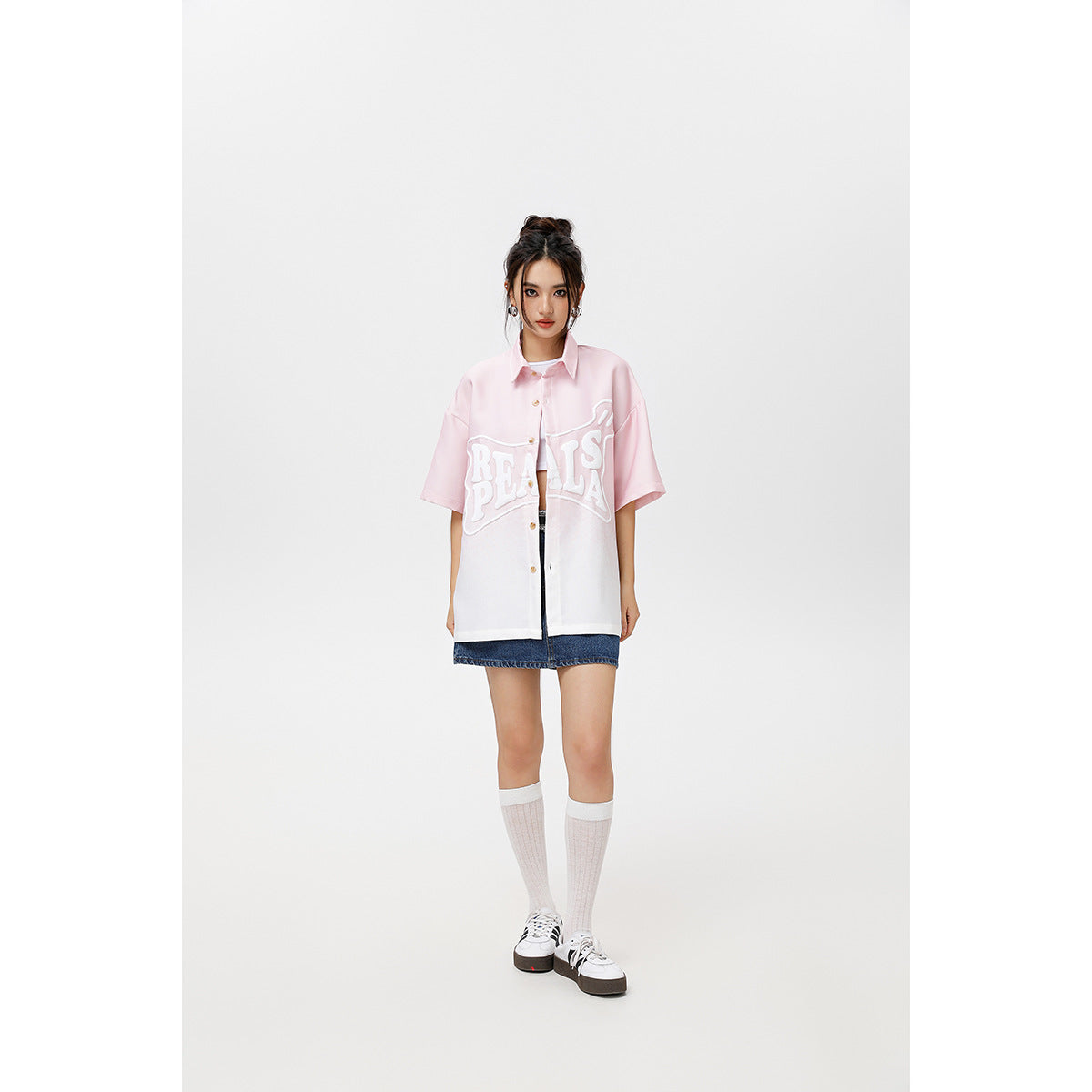 Model wearing an oversized pink-to-white short-sleeve button-up shirt with raised white lettering, styled with a denim skirt.