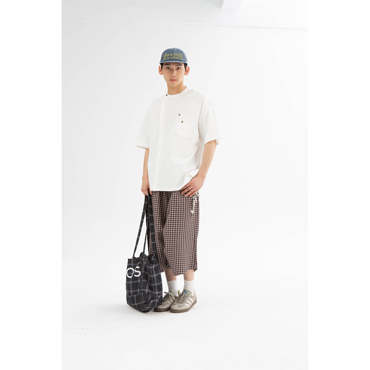 Model wearing a white short-sleeve shirt with a buttoned chest pocket and brown plaid shorts, holding a dark plaid tote.