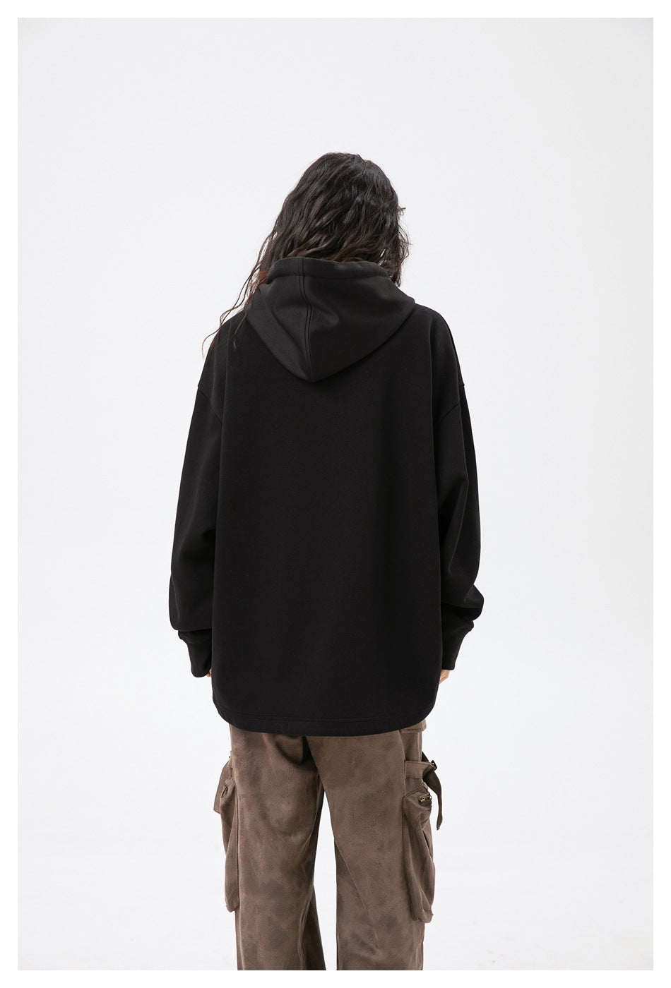 Rear view of a model wearing a plain black oversized hoodie with a hood, paired with brown cargo pants.