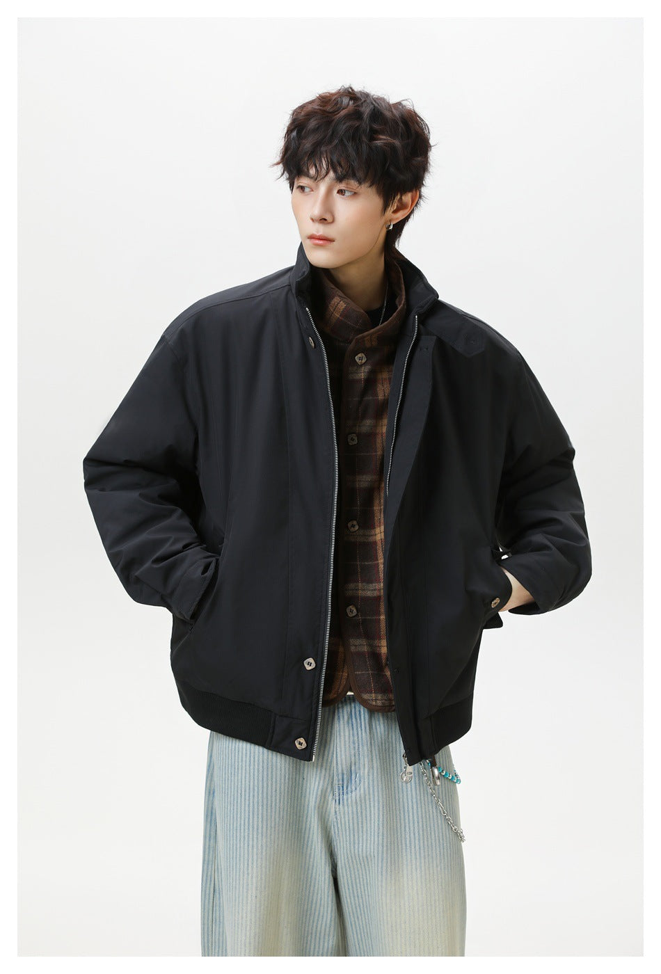 Model wearing an open black jacket with a raised collar over a brown plaid layer and pale blue striped trousers.