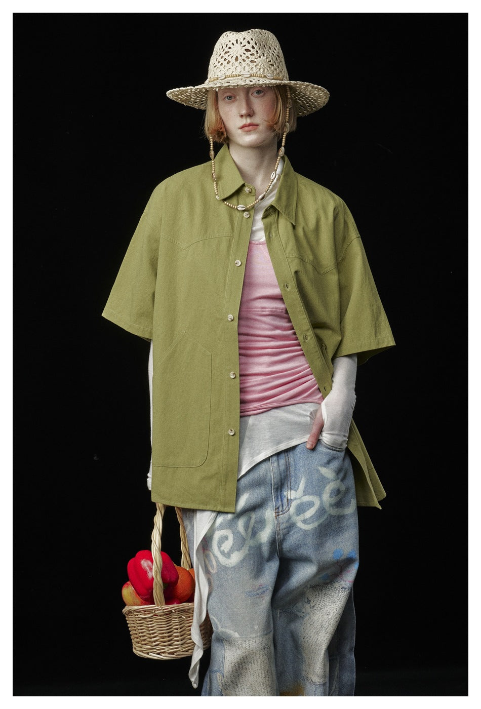 Model wearing an olive-green button-up shirt over a pink top, loose graphic jeans, and a woven sun hat.
