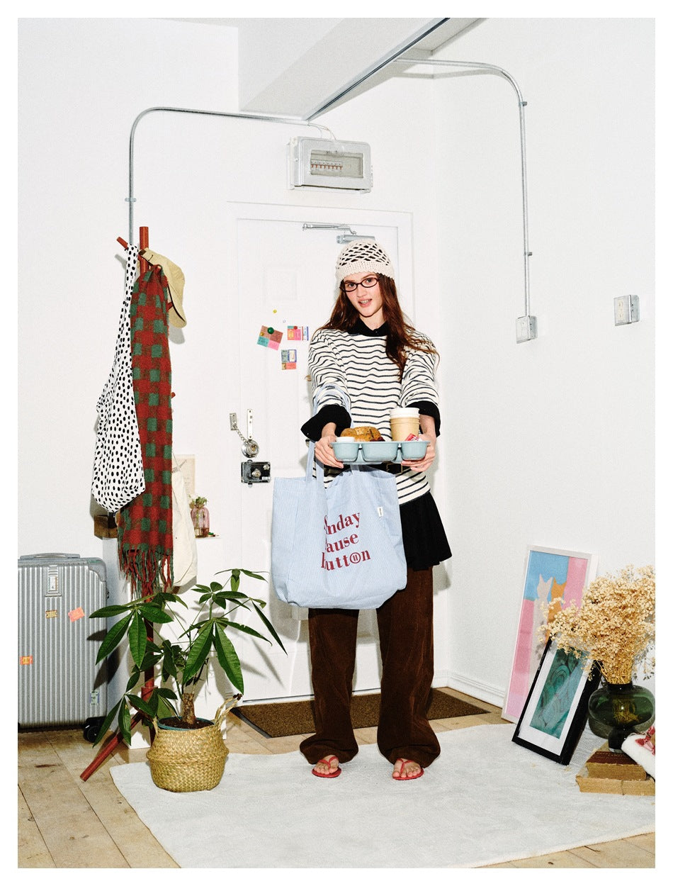 Model wearing a striped long-sleeve top over a black layer with brown trousers, holding a tray and a tote bag