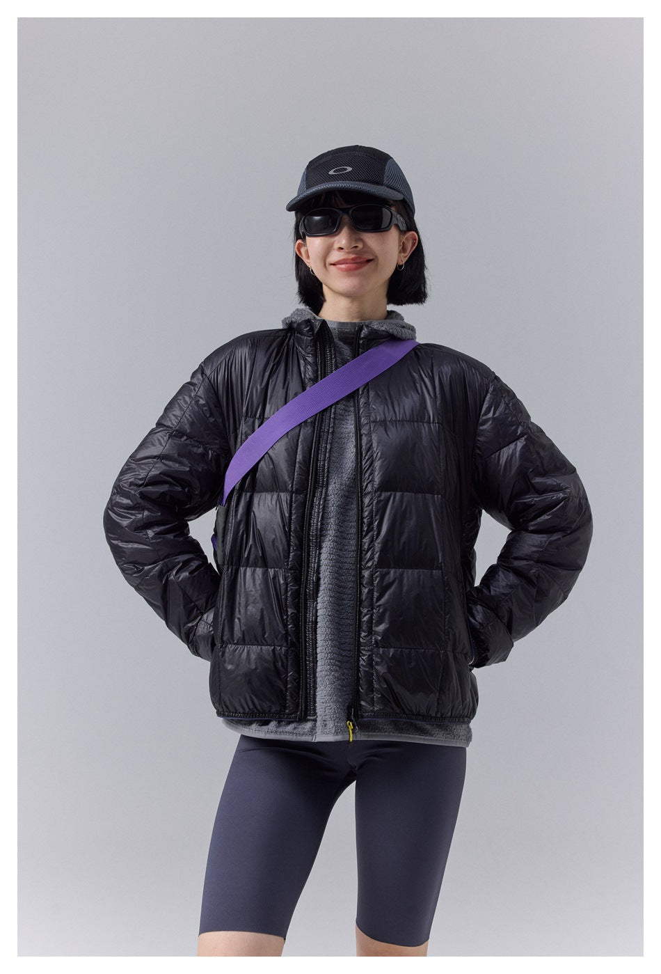 Front view of a model wearing a black quilted jacket over a gray hooded top and dark shorts.