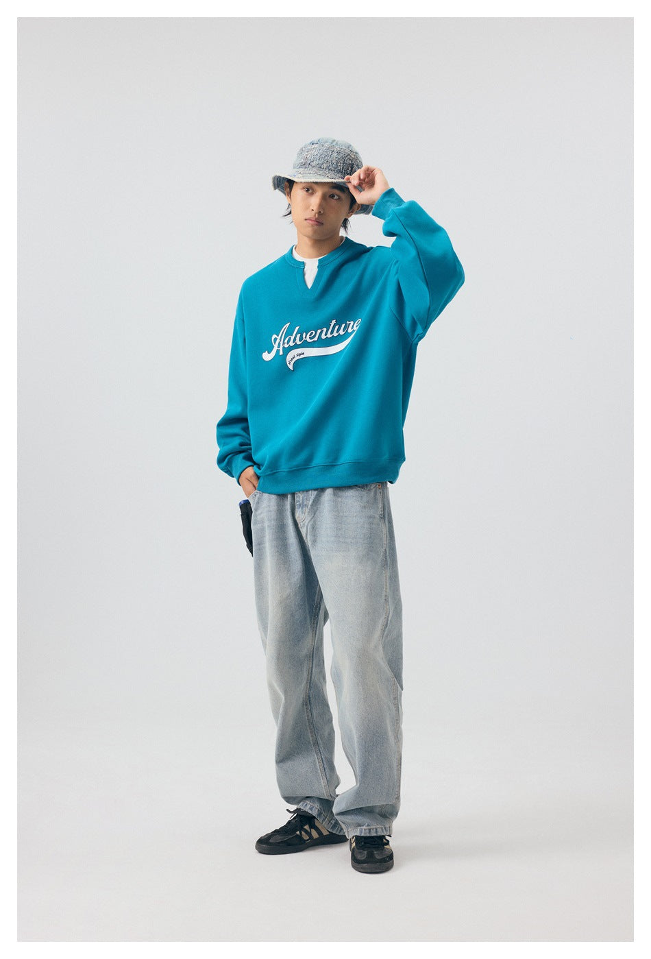 Model in blue Adventure sweatshirt and light-wash jeans, one hand raised to a denim bucket hat