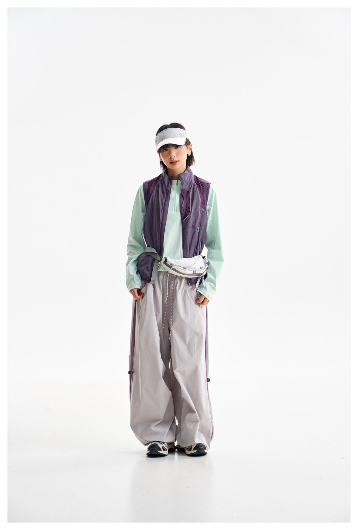 Full-length front view of a woman in a plum utility vest, mint top, loose gray pants, white visor and waist pack.