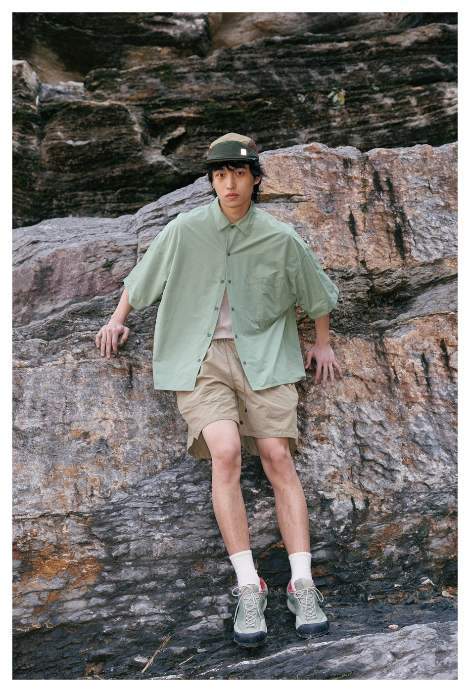 Model wearing a pale sage oversized short-sleeve button-up shirt and tan shorts against a rocky backdrop.