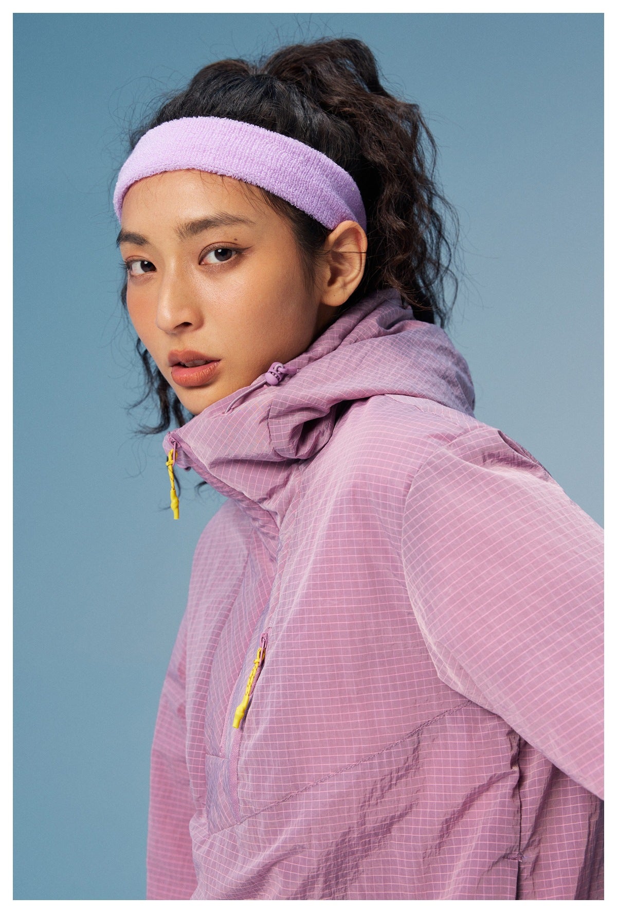 Close-up of a model wearing a lilac grid-pattern hooded pullover with bright yellow zipper pulls and a lavender headband.