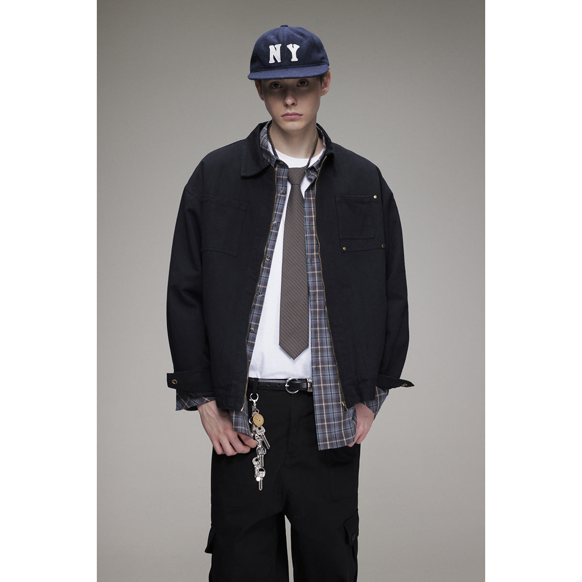 Model wearing a black utility jacket over a plaid shirt, white tee and tie, with black cargo pants and a navy cap.