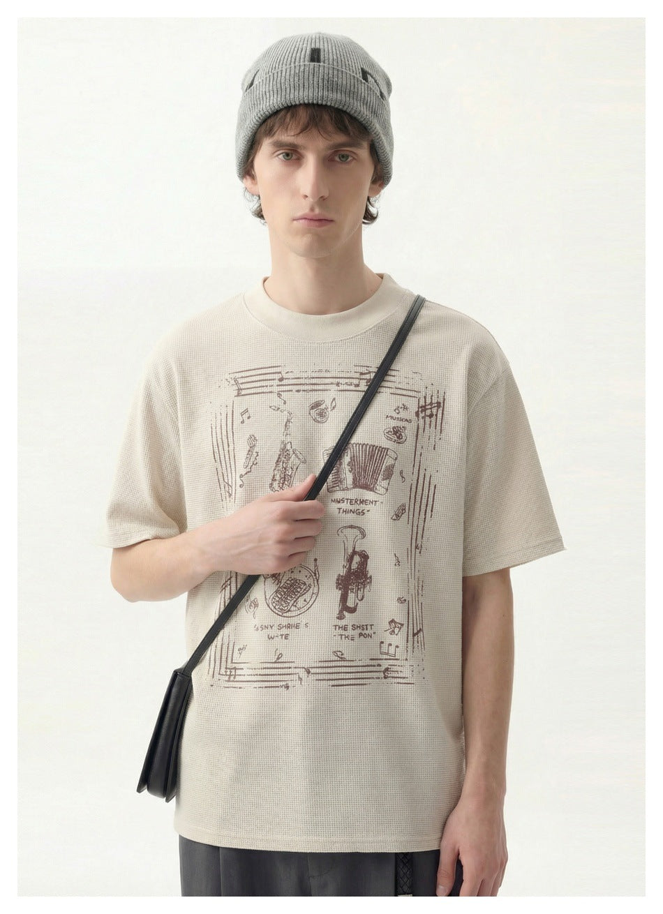 Front view of a model wearing an ivory T-shirt with a large brown music-themed graphic and a gray beanie.