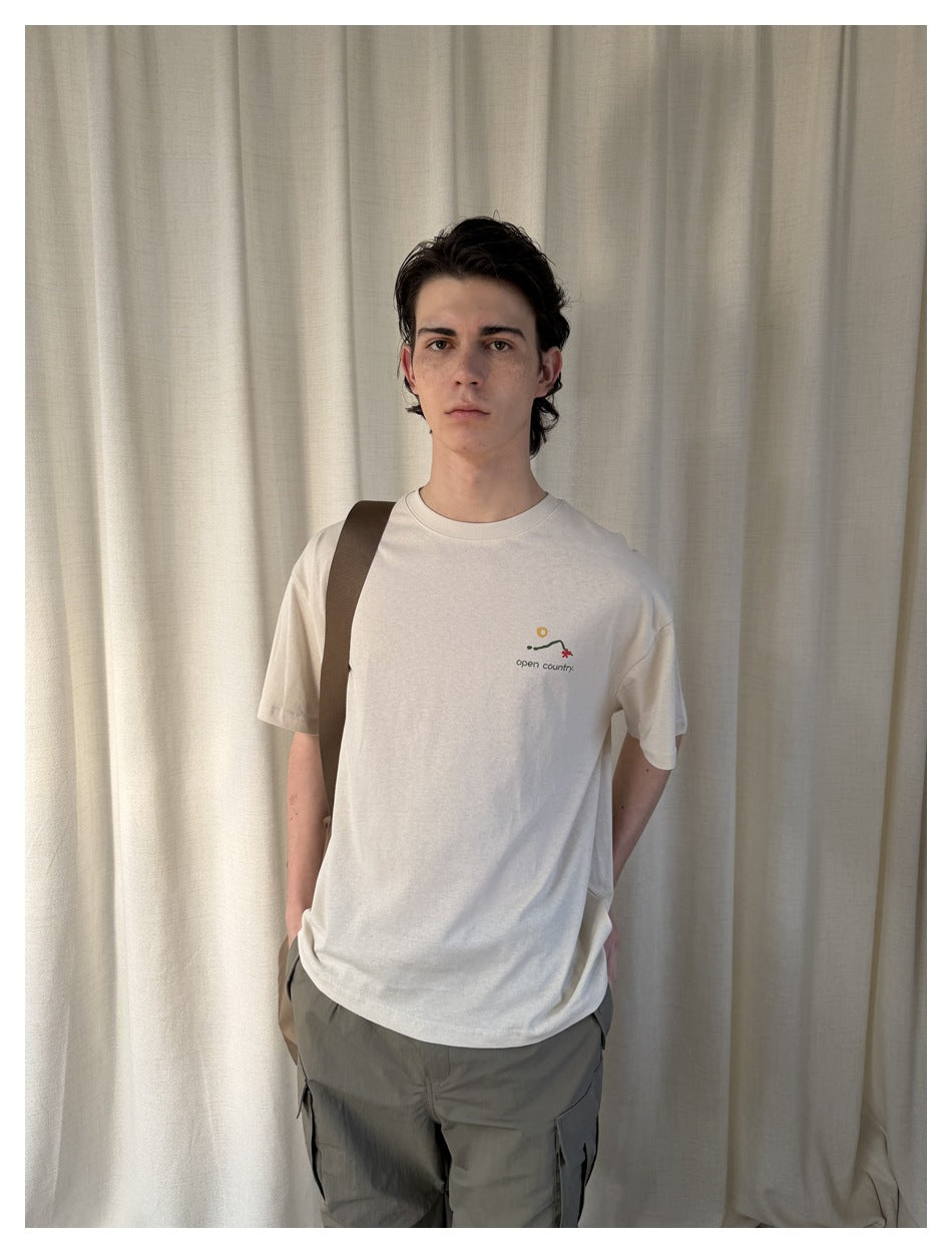Man wearing a beige relaxed t-shirt with small chest print, a brown shoulder strap and gray cargo pants against a curtain
