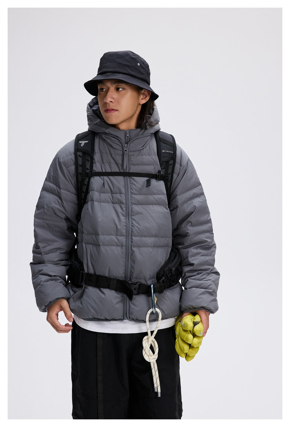 Close view of a model in a gray hooded puffer jacket with a backpack, bucket hat and yellow gloves